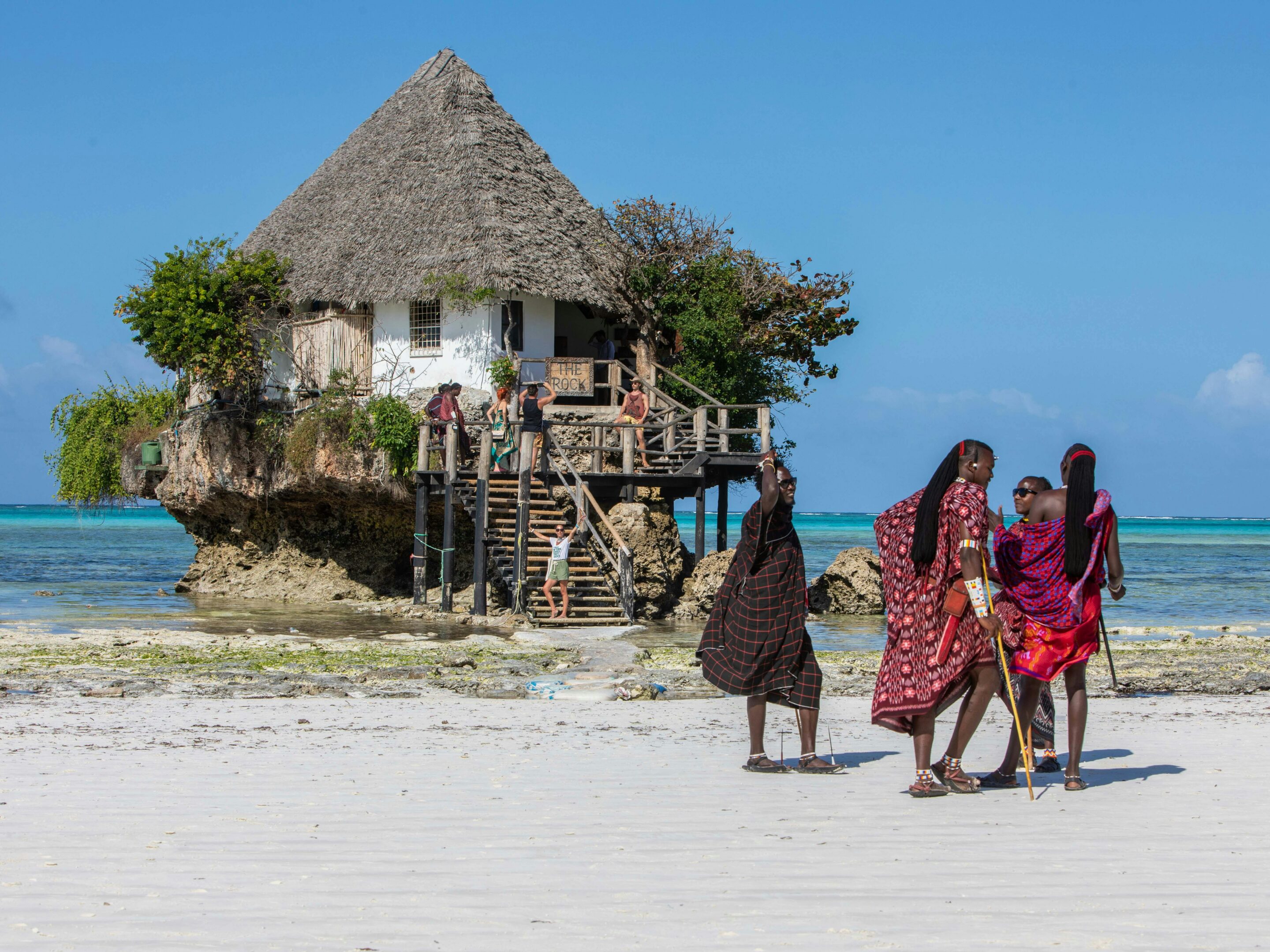 A thatched-roof restaurant sits atop a rock in shallow turquoise water off Zanzibar. Four people in colorful traditional clothing walk along the sandy beach in the foreground beneath the clear blue sky of the Spice Islands.