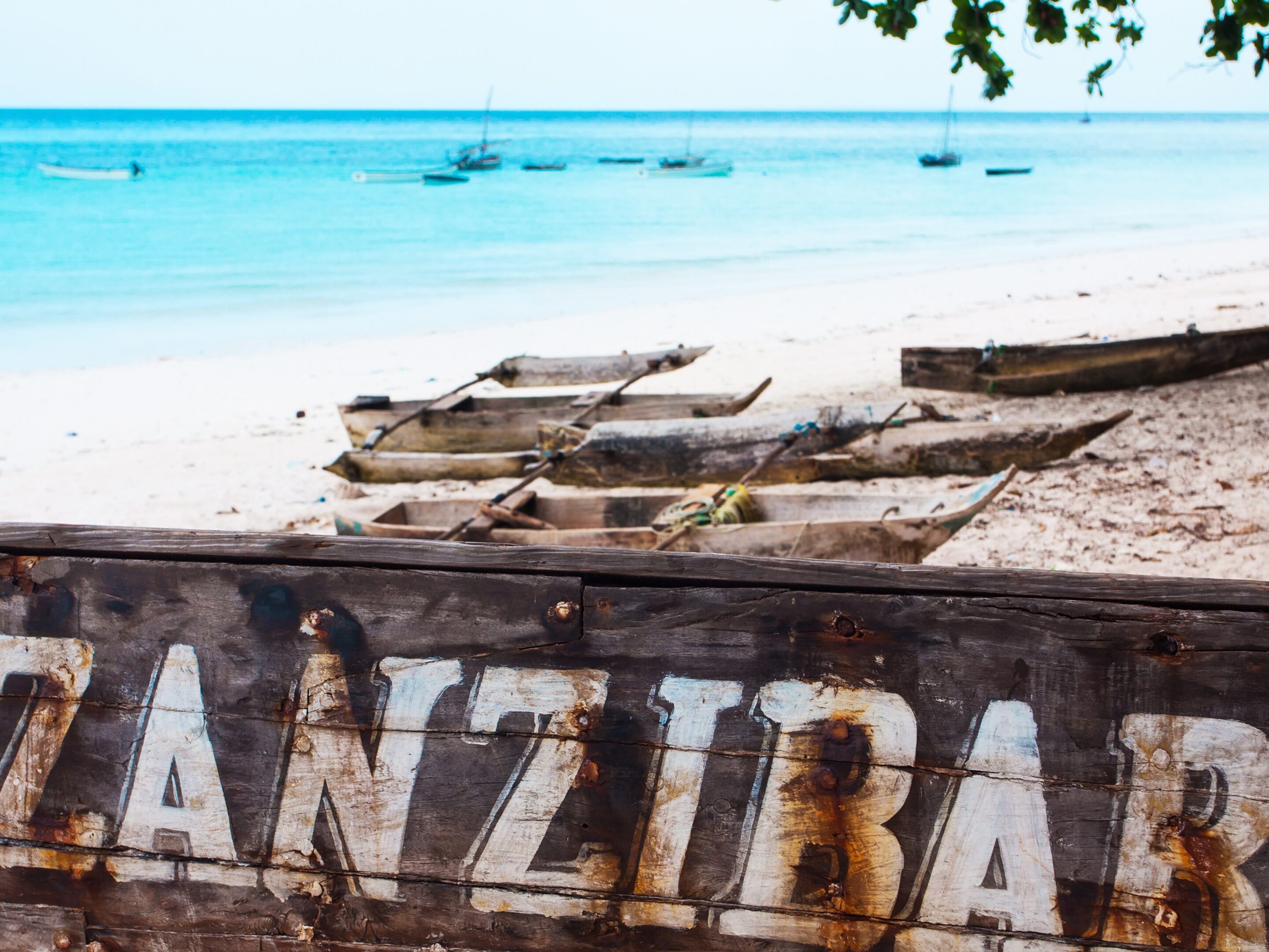 A weathered wooden sign reading ZANZIBAR sits on a sandy beach in the Spice Islands, with wooden boats and turquoise ocean waters in the background, under a partly cloudy sky.