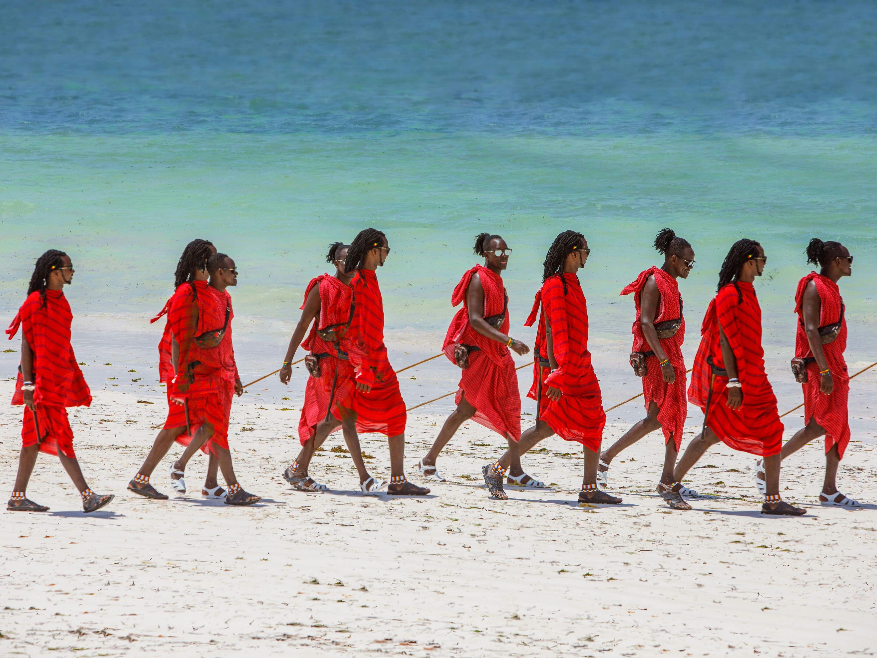 A group of people wearing bright red traditional attire walk in a line along a sandy Zanzibar beach, with turquoise ocean water in the background.