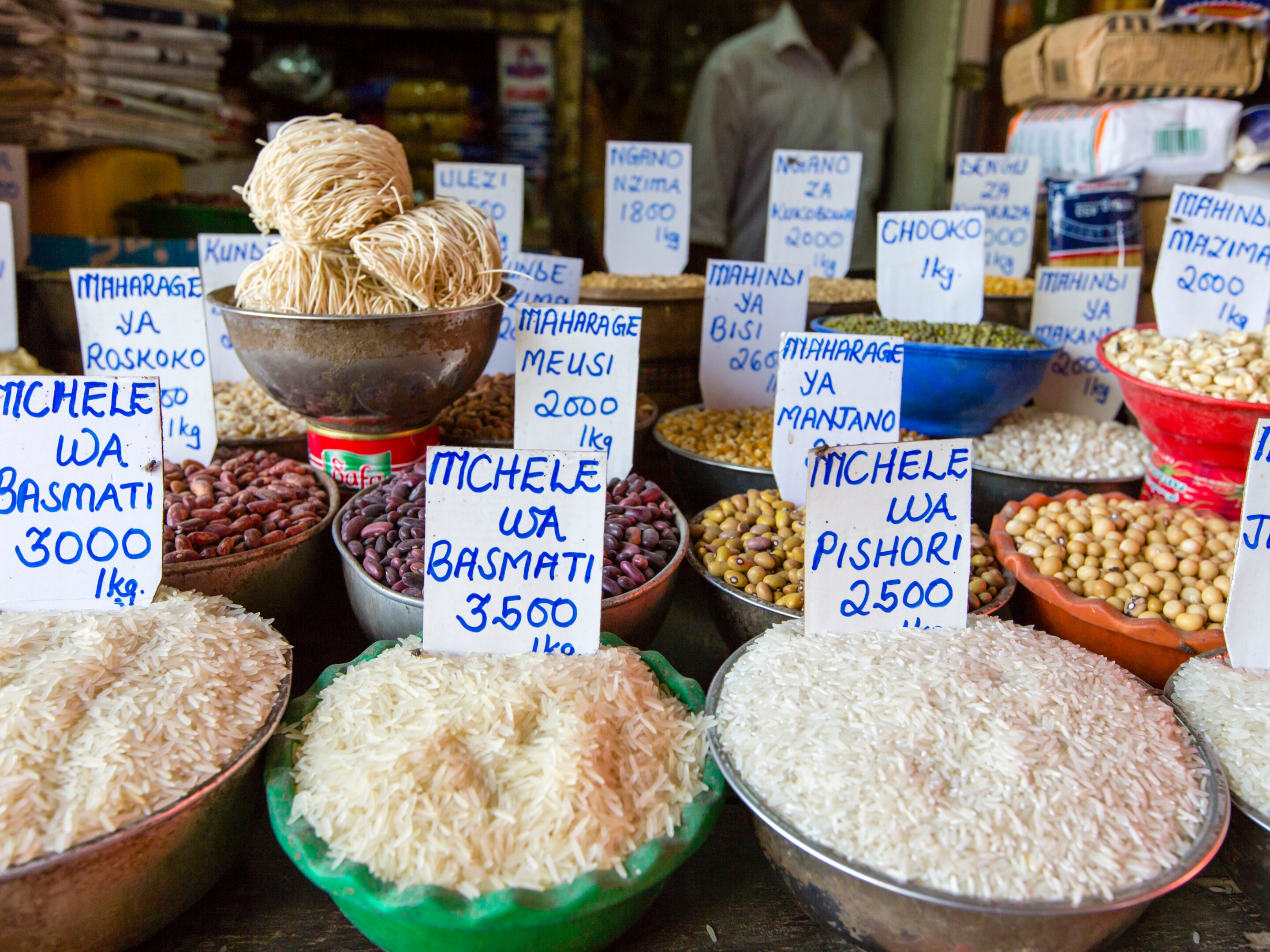 Various bowls of rice, beans, noodles, and grains with handwritten price signs in Swahili are displayed at a Zanzibar market stall. The colorful ingredients evoke the vibrant atmosphere of the Spice Islands.