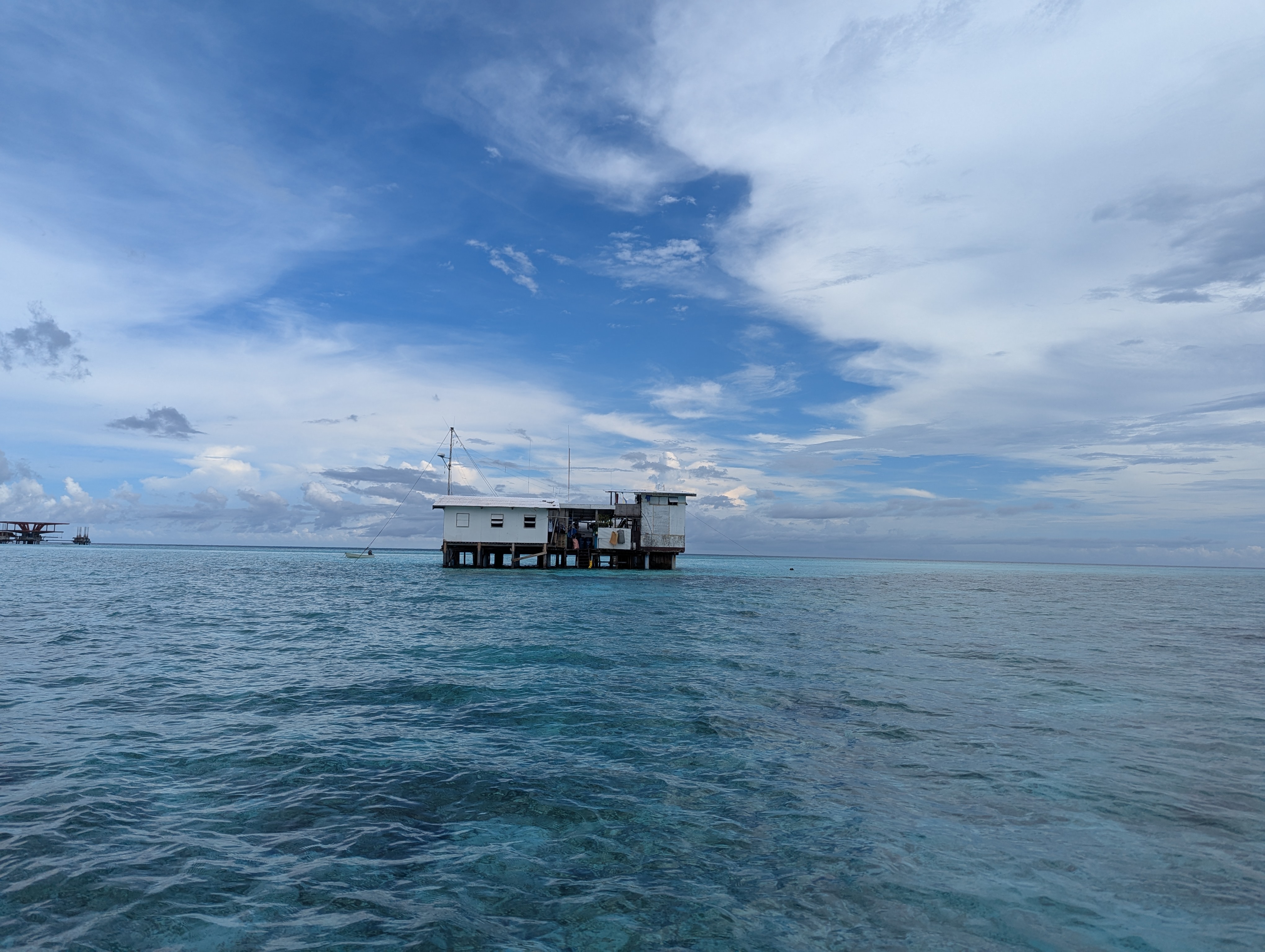 A small, white stilt house sits above clear, shallow ocean water in the Wild Philippines under a cloudy blue sky.