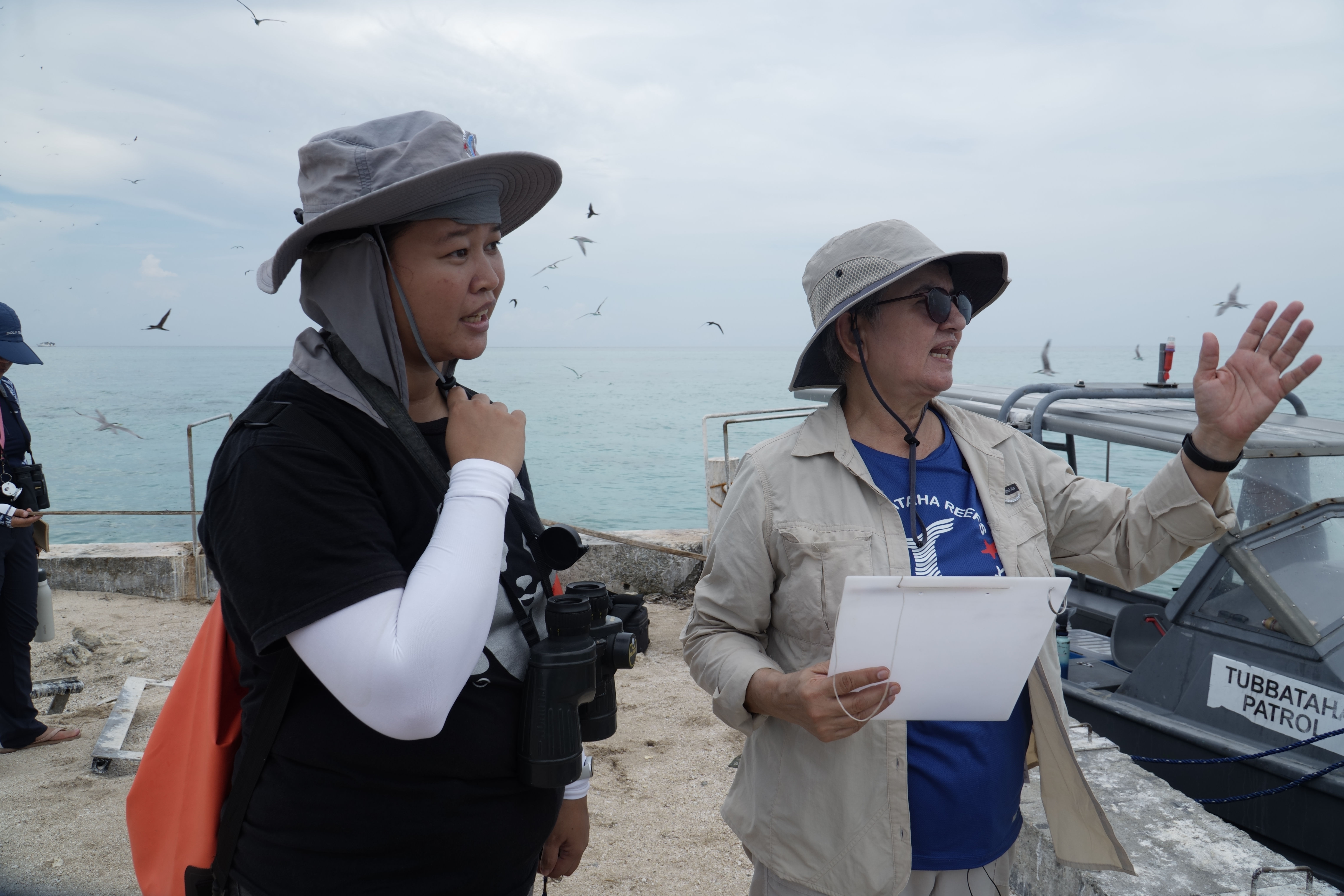 Two people in sun hats stand on a pier, one holding binoculars and the other gesturing toward the Wild Philippines ocean while holding a clipboard.