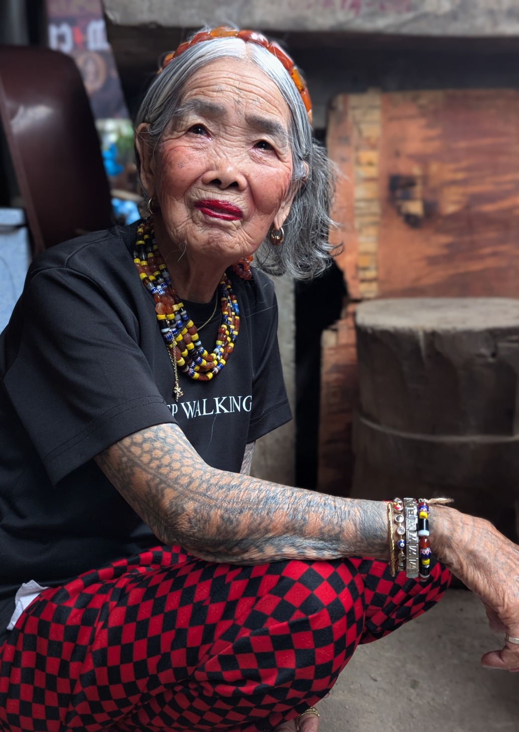 An elderly woman with grey hair, a beaded headband, and necklaces, wearing a black t-shirt and red and black checkered pants, showing extensive arm tattoos as a guardian of nature in the Wild Philippines.